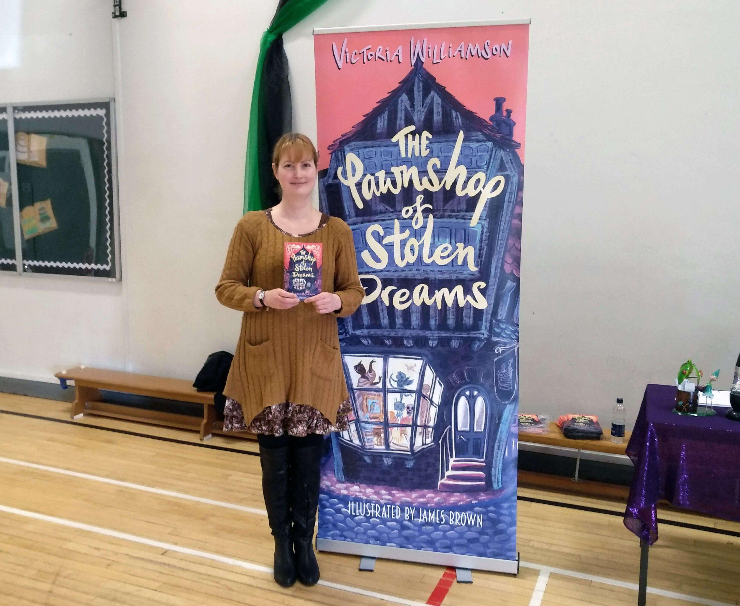 The Pawnshop of Stolen Dreams author Victoria Williamson holds her book next to a large banner promoting her book.